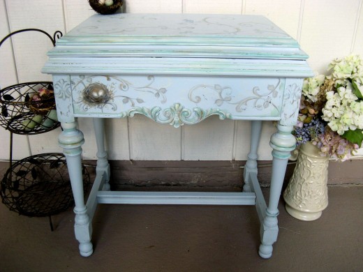 This charmingly distressed "shabby chic" console table is an old sewing cabinet in disguise, hand painted in layered blue tones with verdigris patina applied to the scrollwork, with a delicate hand-painted birds nest as accent.