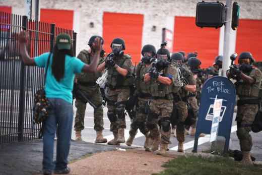 A black man holds up hands in fear, facing several heavily armed police who are pointing the barrels of their guns at him.