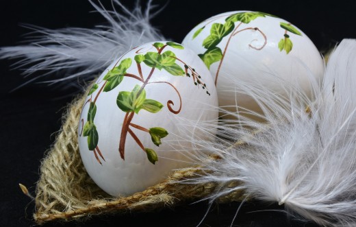 Two white eggs with painted branches and leaves placed in a basket of feathers. 