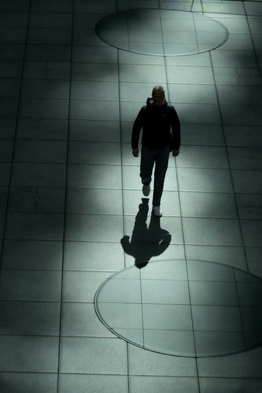 A man in shadow walks alone through pools of light on a tiled floor, photo by Sue Winston on Unsplash