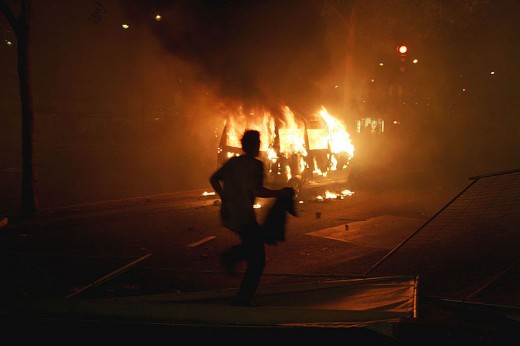 A man runs over a fence in front of a van set ablaze by a group of rioters.