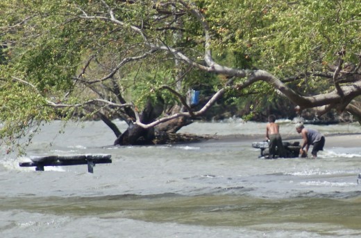 An empty washing table and two other women washing clothes along the shoreline.