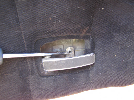 A screwdriver points to the screw that holds the door handle back plate on. (The back plate has been removed in this photo.) 