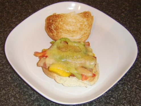 Cheddar cheese is melted over bacon and bell peppers on a toasted bread roll