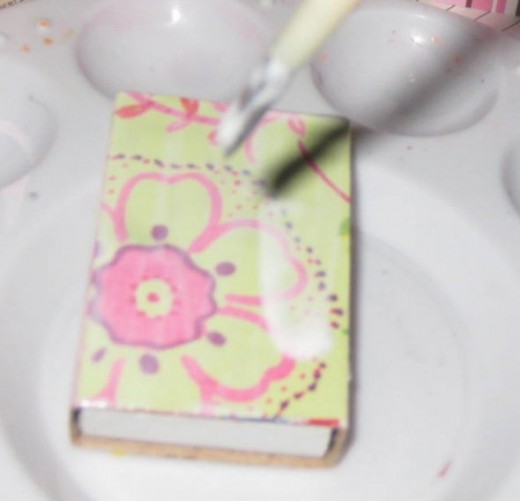 Use decoupage medium like Mod Podge to adhere the paper to the matchbox. Do one side at a time letting the Mod Podge dry completely before adding paper to the other side.