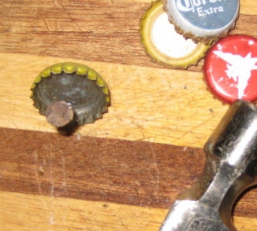 There are several ways to punch a hole in a bottle cap. One way is to use a nail and hammer. Place the nail on the inside of the bottle cap on the rim's flat edge (flat as it sits on a piece of wood). Hold the nail and hit the nail (not your finge