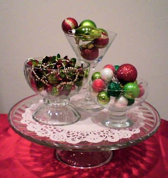 Martini glass and 1 small bowl filled with ornaments, larger bowl filled with garland, placed on a doily on the cake stand.